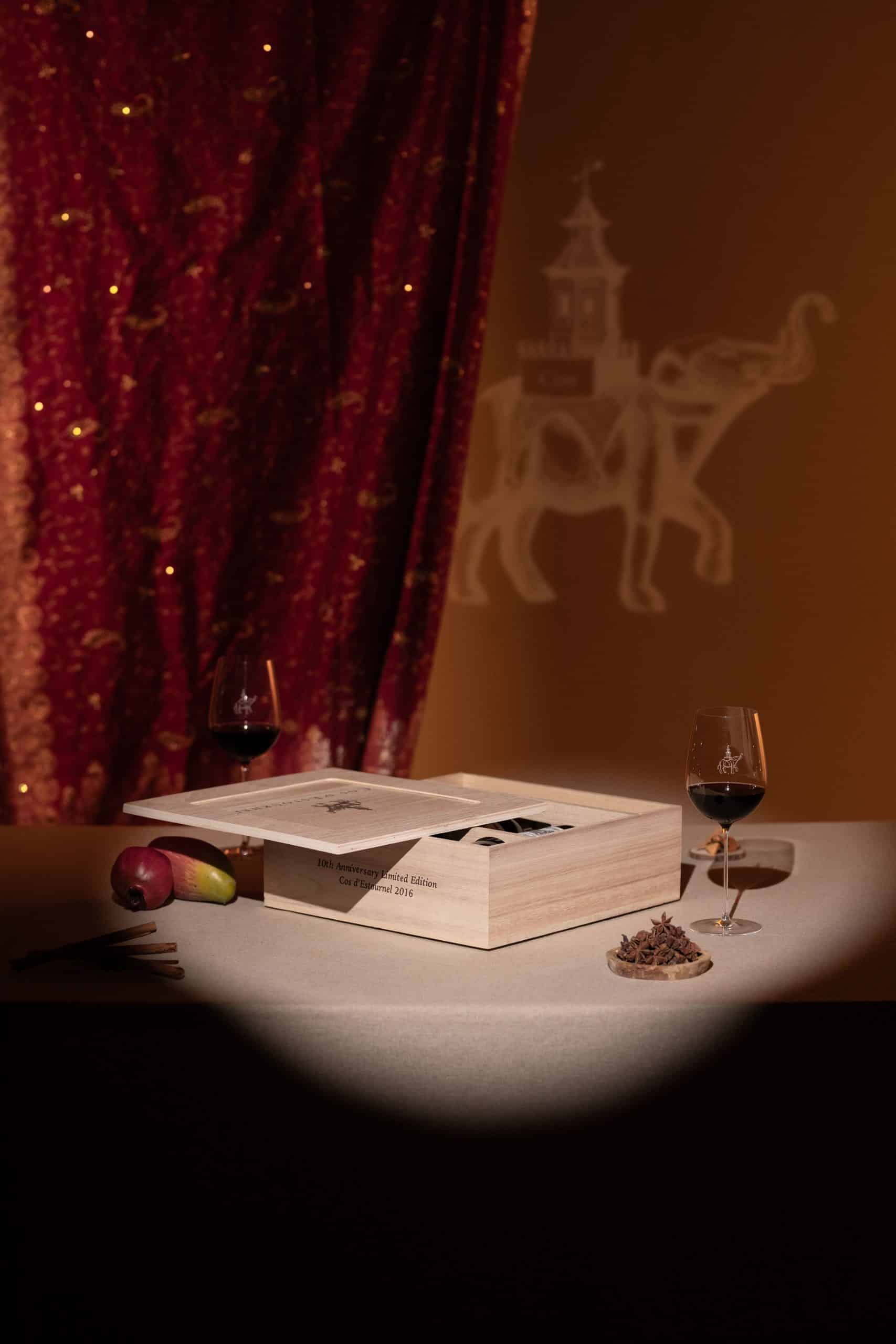 Still life scene: a wooden box on a table with a menu, two glasses of red wine, apples, cinnamon sticks, and star anise, against a red velvet curtain and a pale horse-drawn carriage projection on the wall.